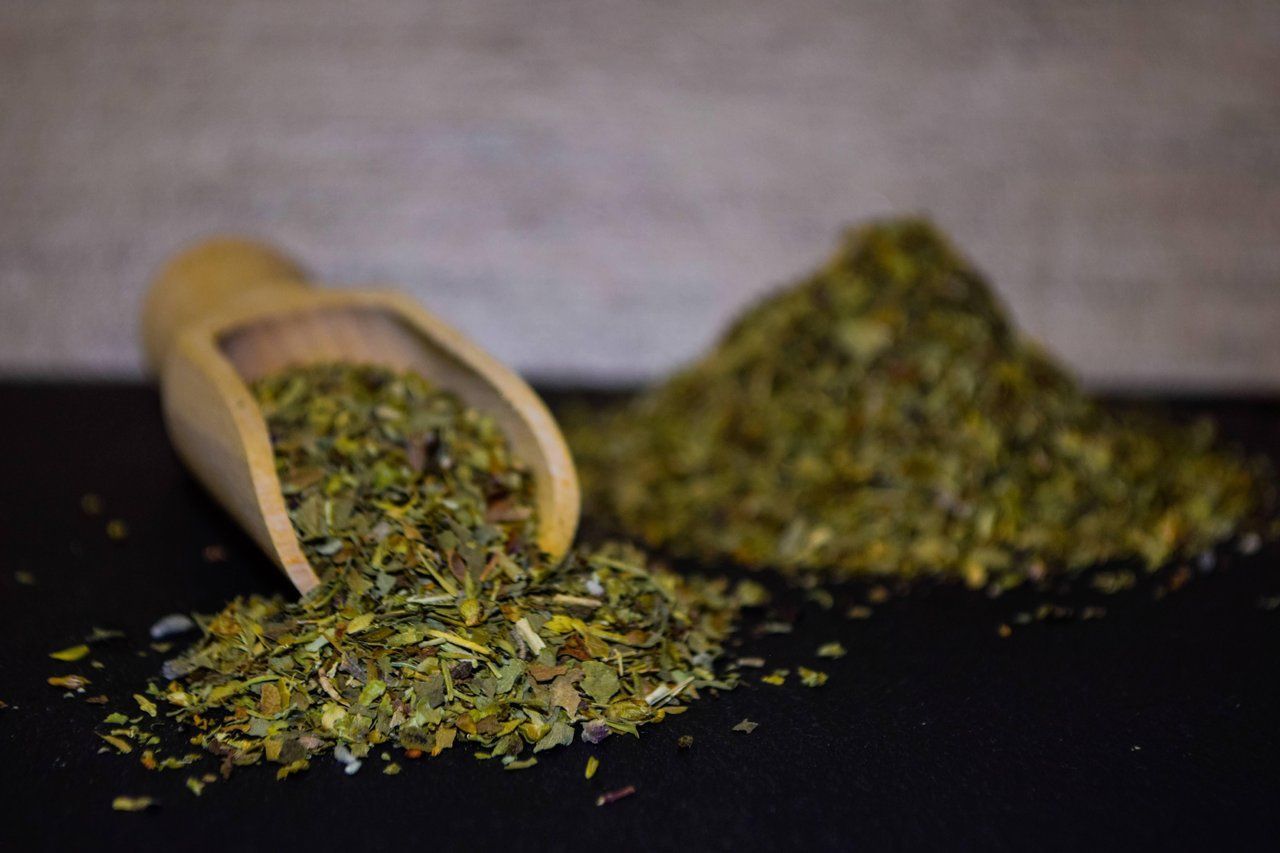 Eine Holzschaufel schüttet getrocknete grüne Kräuter auf eine dunkle Oberfläche.