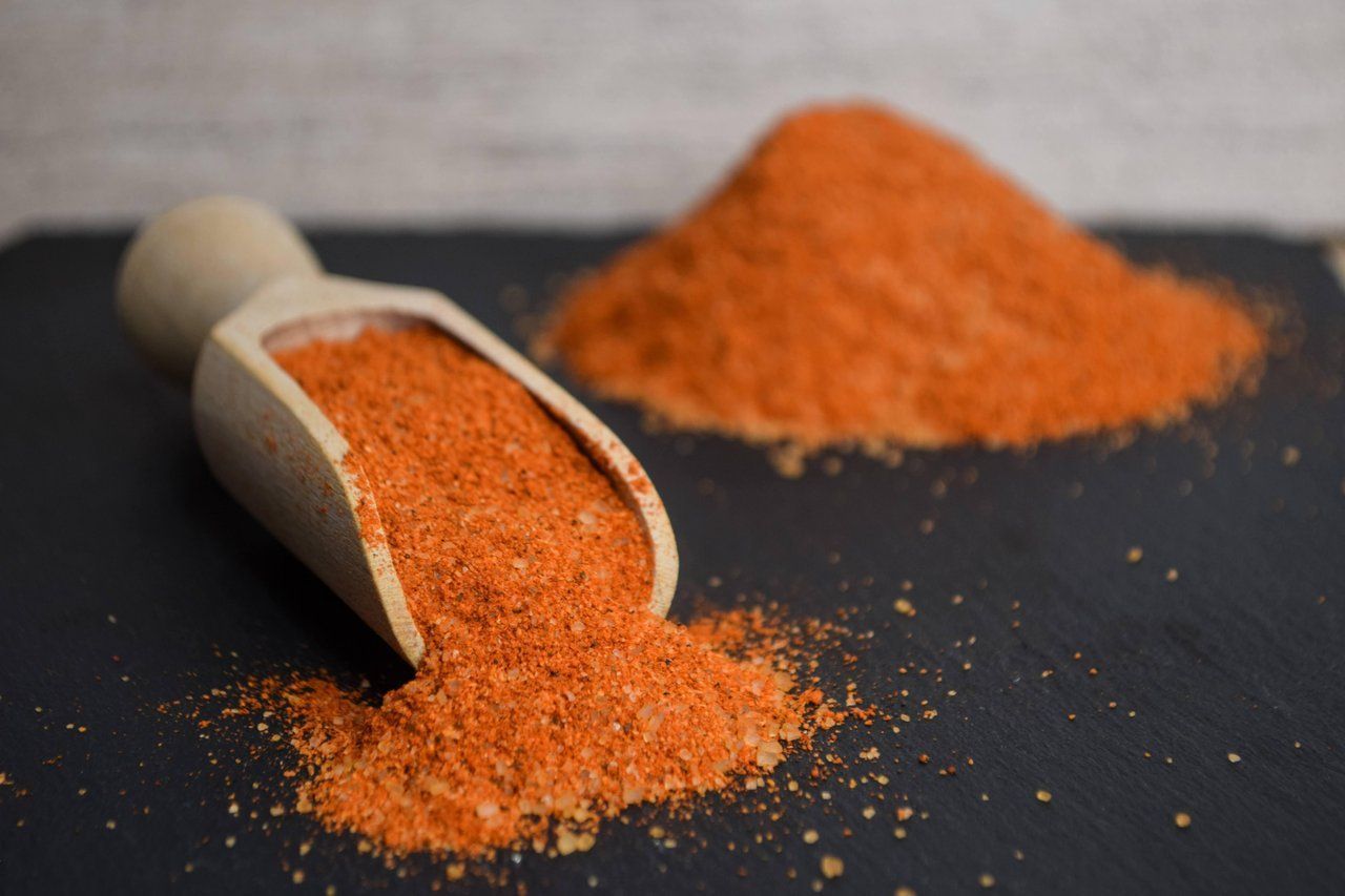 Eine Holzschaufel schüttet orangefarbenes Gewürz auf eine schwarze Fläche; im Hintergrund ist ein kleiner Haufen zu sehen.