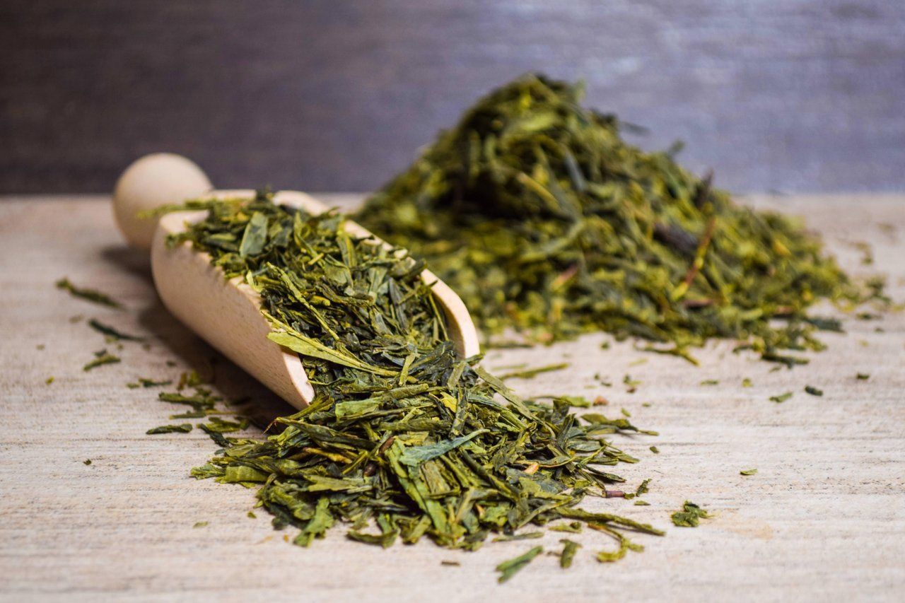 Zwei Holzschaufeln verschütten getrocknete grüne Teeblätter auf einer rustikalen Oberfläche.