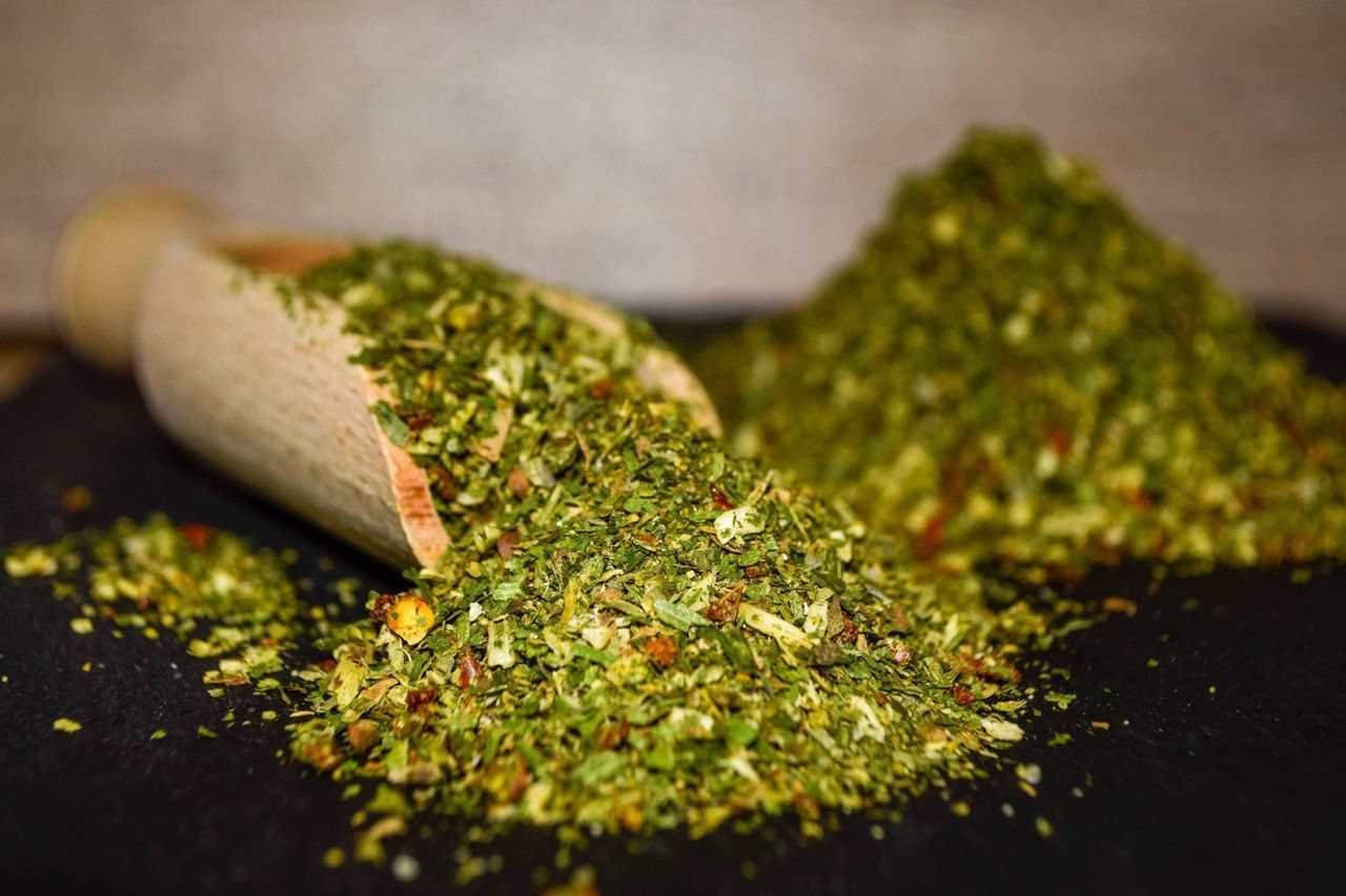 Eine Holzschaufel verschüttet grüne und rote getrocknete Kräuter auf einer dunklen Oberfläche.