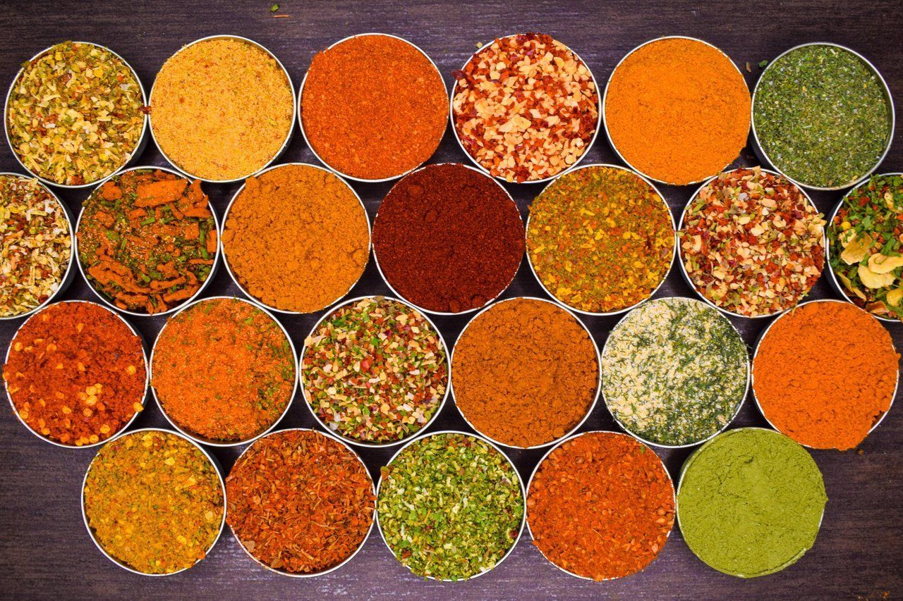 Verschiedene bunte Gewürze und Körner, arrangiert in runden Schalen auf einer dunklen Oberfläche.