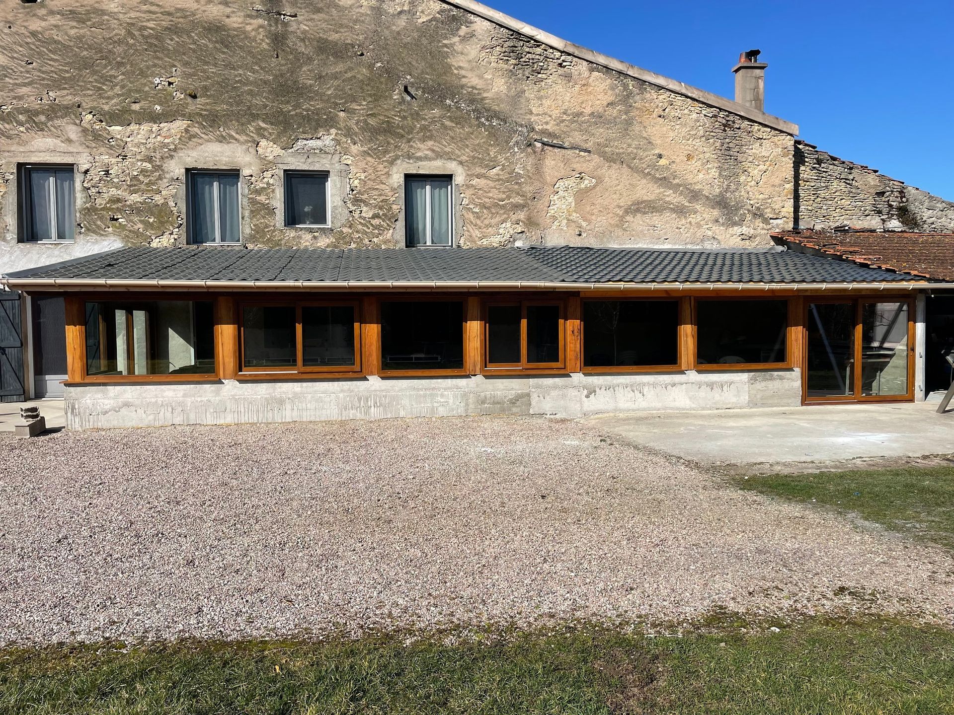 Un bâtiment long et bas, doté de grandes fenêtres et d'une terrasse en gravier, se dresse devant un bâtiment en pierre patinée par le temps.