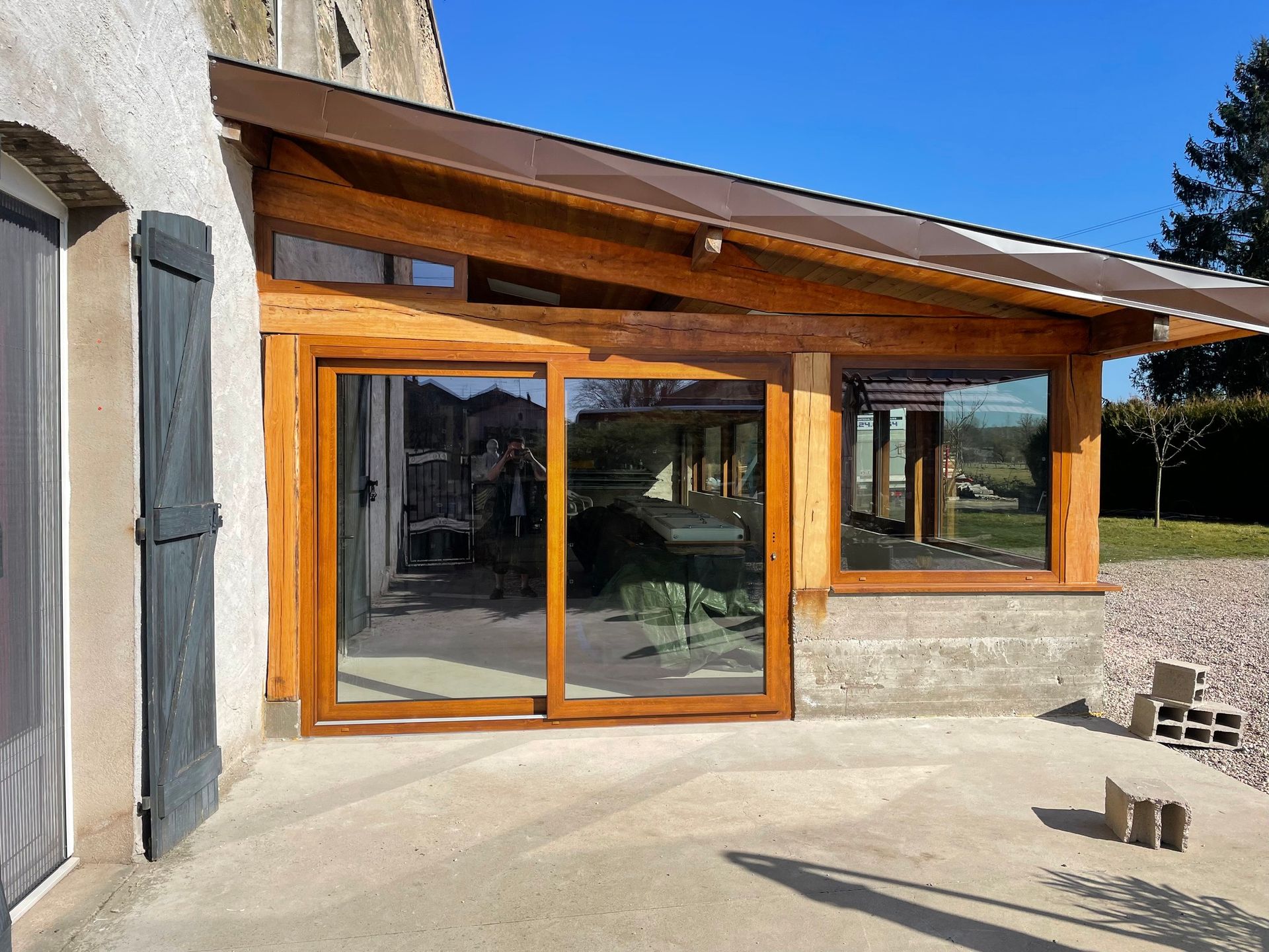 Patio à ossature bois avec portes coulissantes en verre, attenant à un bâtiment clair. Ciel bleu dégagé.