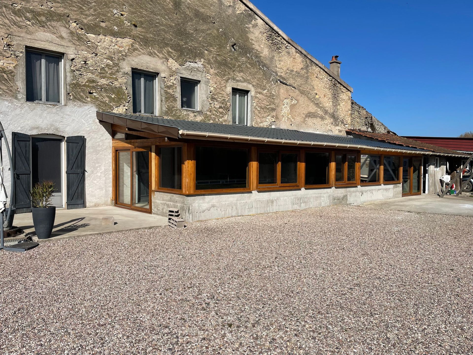 Patio à ossature bois avec portes coulissantes en verre, attenant à un bâtiment clair. Ciel bleu dégagé.