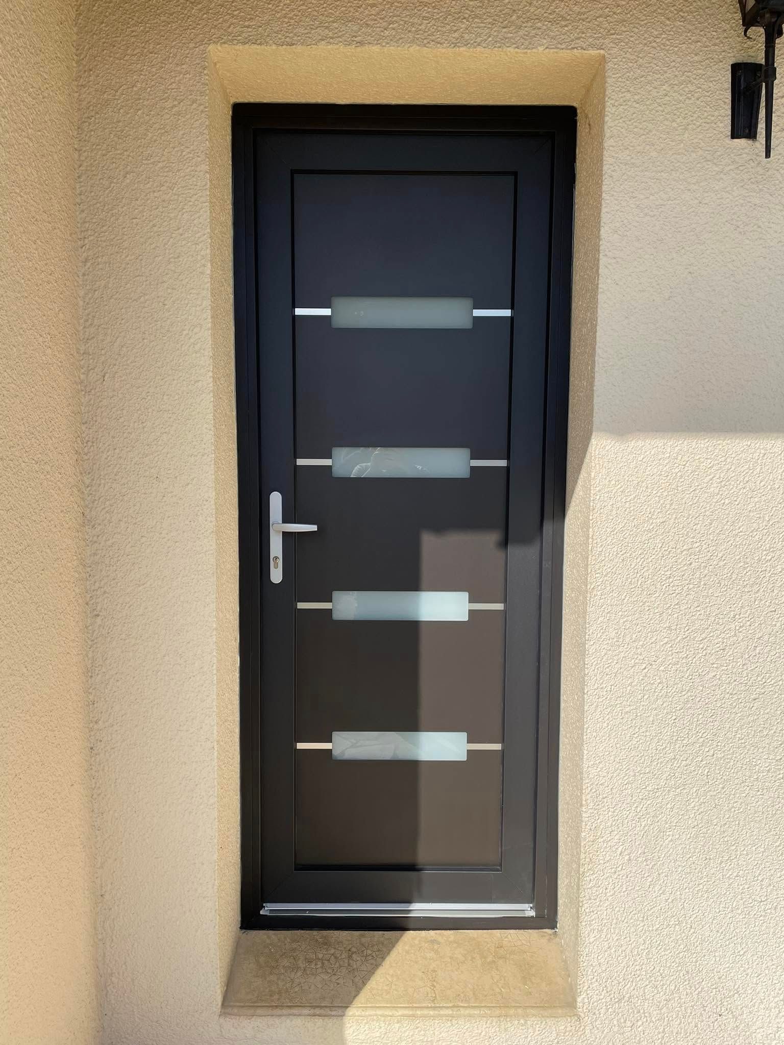 Porte d'entrée noire avec panneaux de verre dépoli, poignée argentée, encastrée dans un mur beige clair.