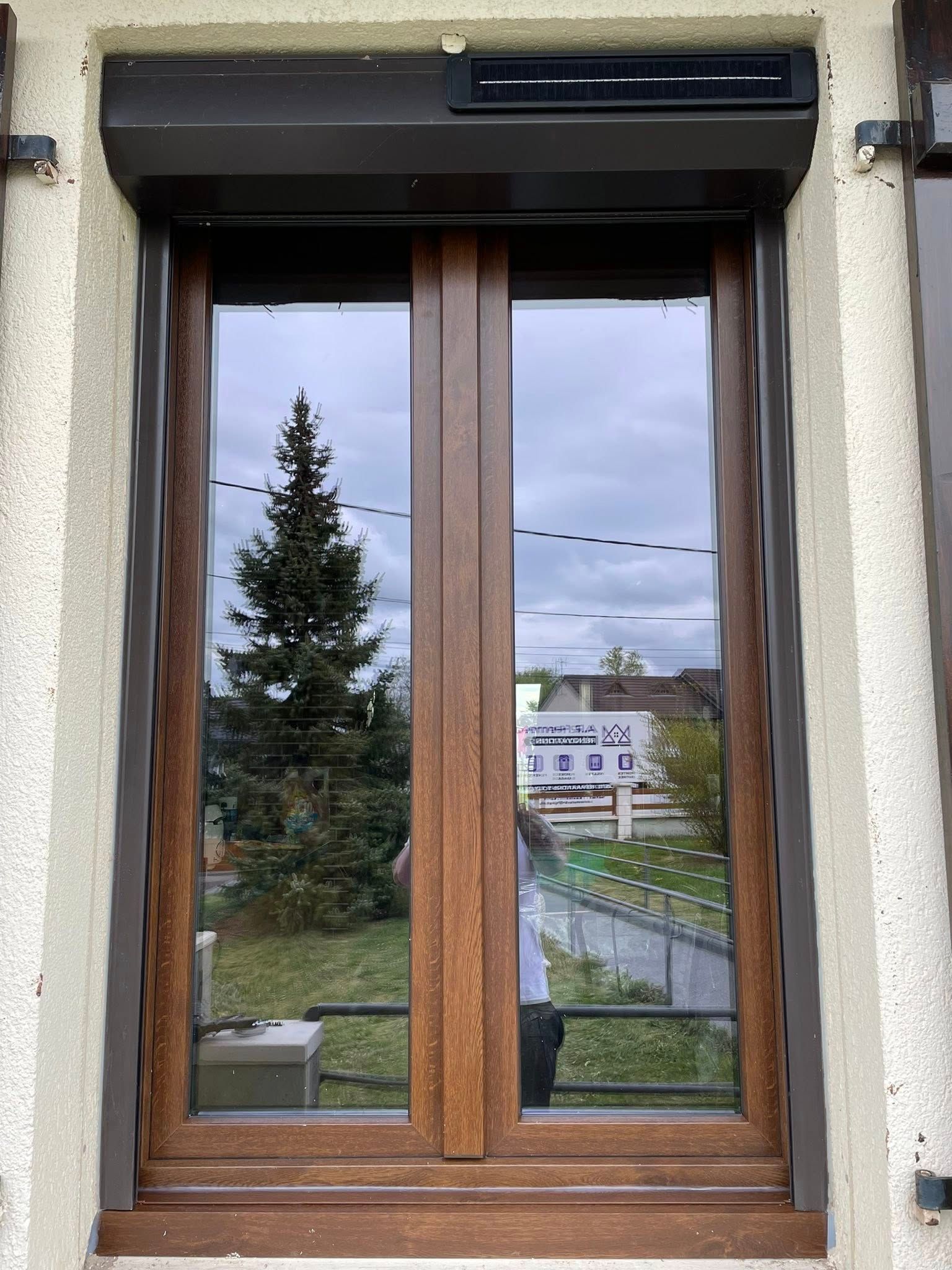 Fenêtre à cadre brun avec verre réfléchissant, volet brun foncé, donnant sur des arbres et un ciel nuageux.