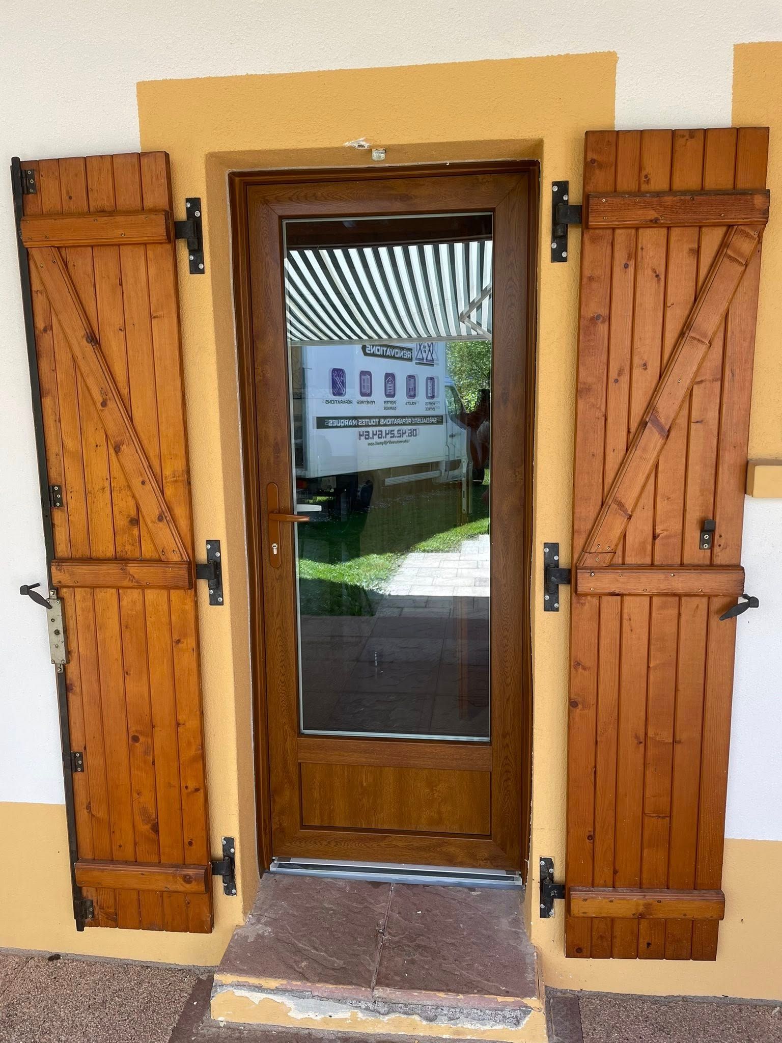 Porte en bois brun avec panneau de verre, flanquée de volets en bois, sur un bâtiment jaune et beige.