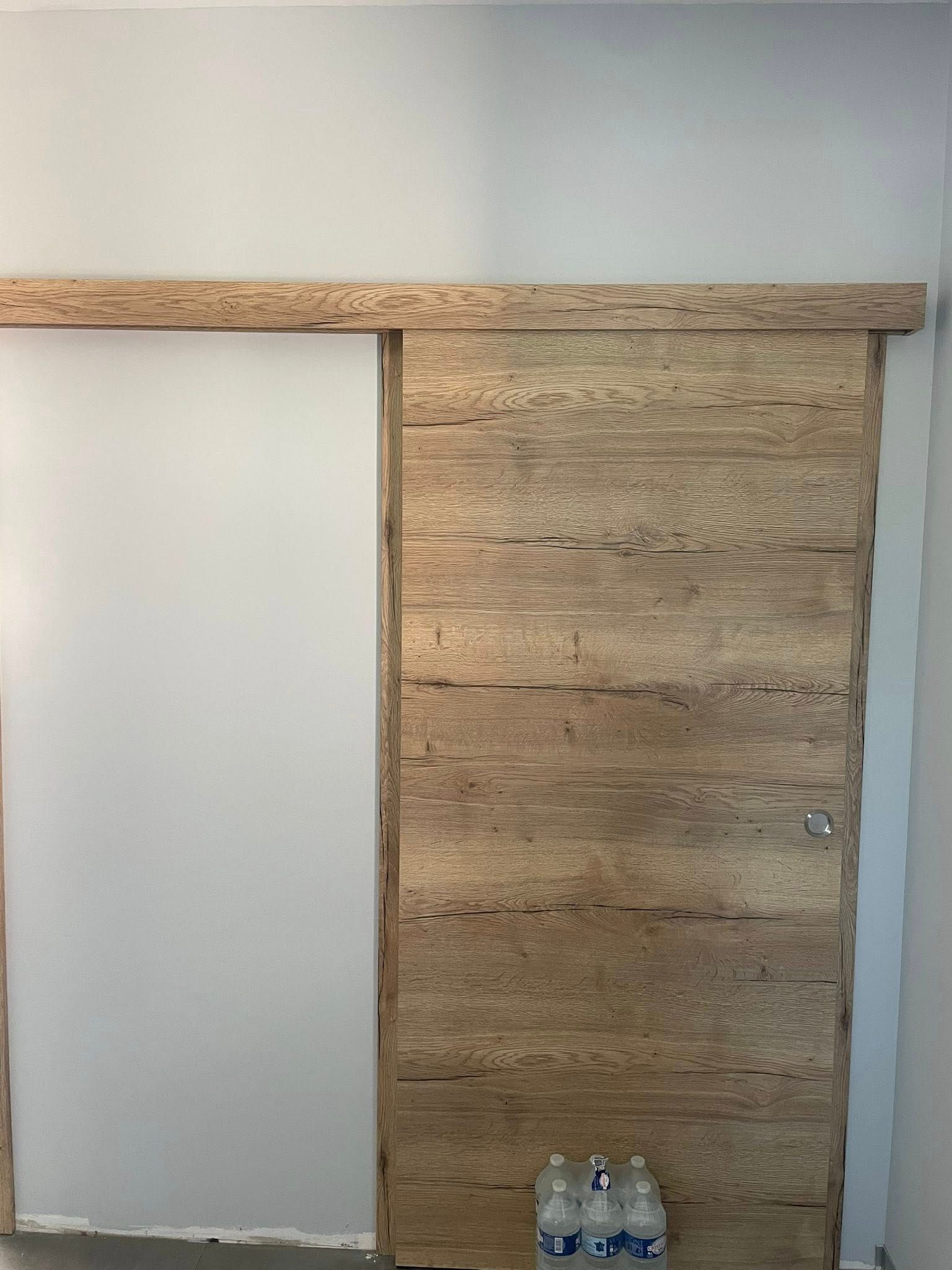 Porte coulissante en bois, entrouverte. Bois clair veiné, montée sur un rail. Trois bouteilles d'eau sont posées au fond.