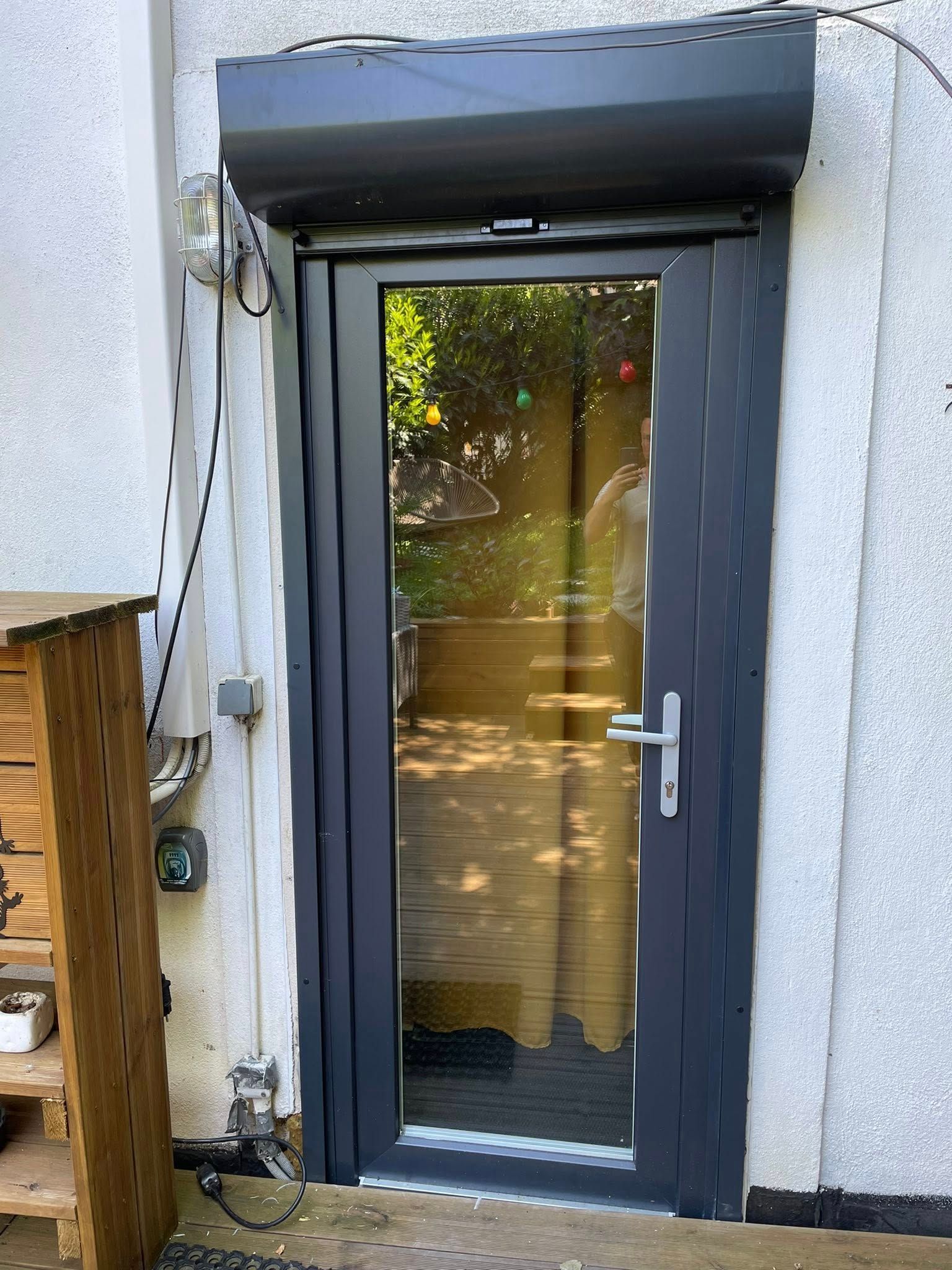 Porte grise vitrée avec volet roulant et poignée argentée. À l'extérieur, une terrasse en bois.