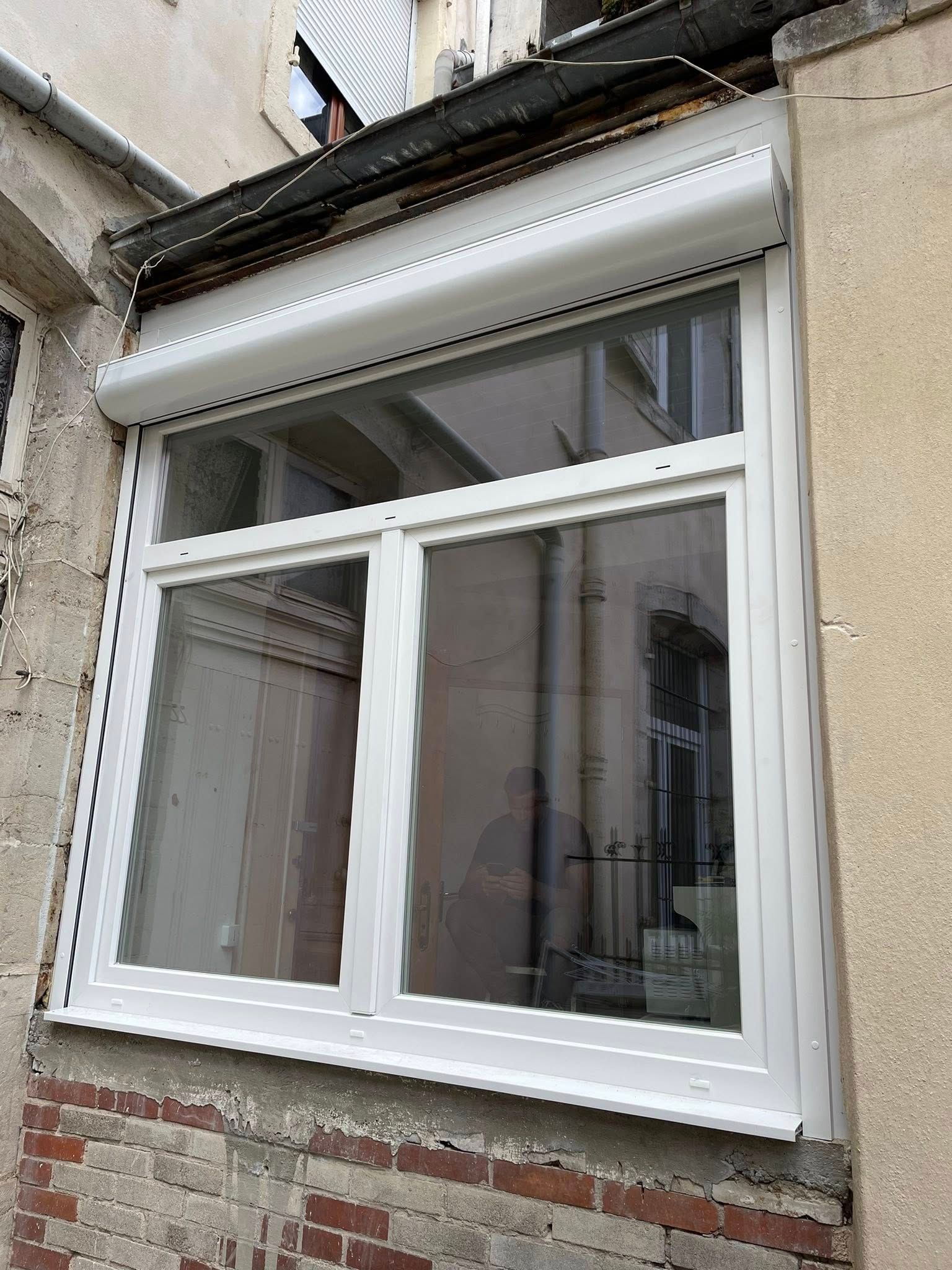 Fenêtre à cadre blanc avec volet roulant blanc fermé, sur la façade d'un bâtiment en briques et en stuc.