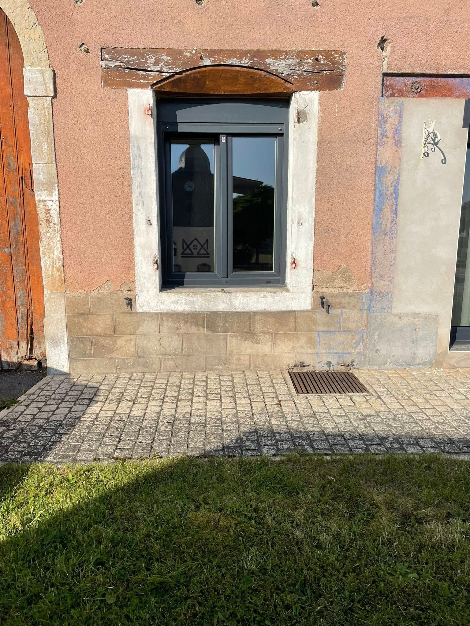 Fenêtre dans un immeuble rose avec poutre en bois au-dessus. Cadre de fenêtre gris, chemin pavé et herbe.