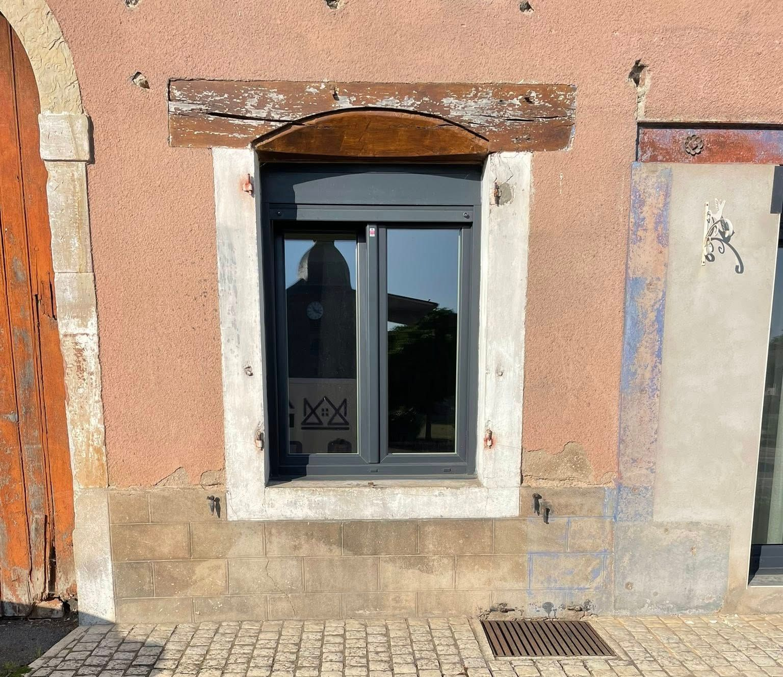 Fenêtre dans un mur en stuc rose, avec un cadre gris foncé, sous une poutre en bois.