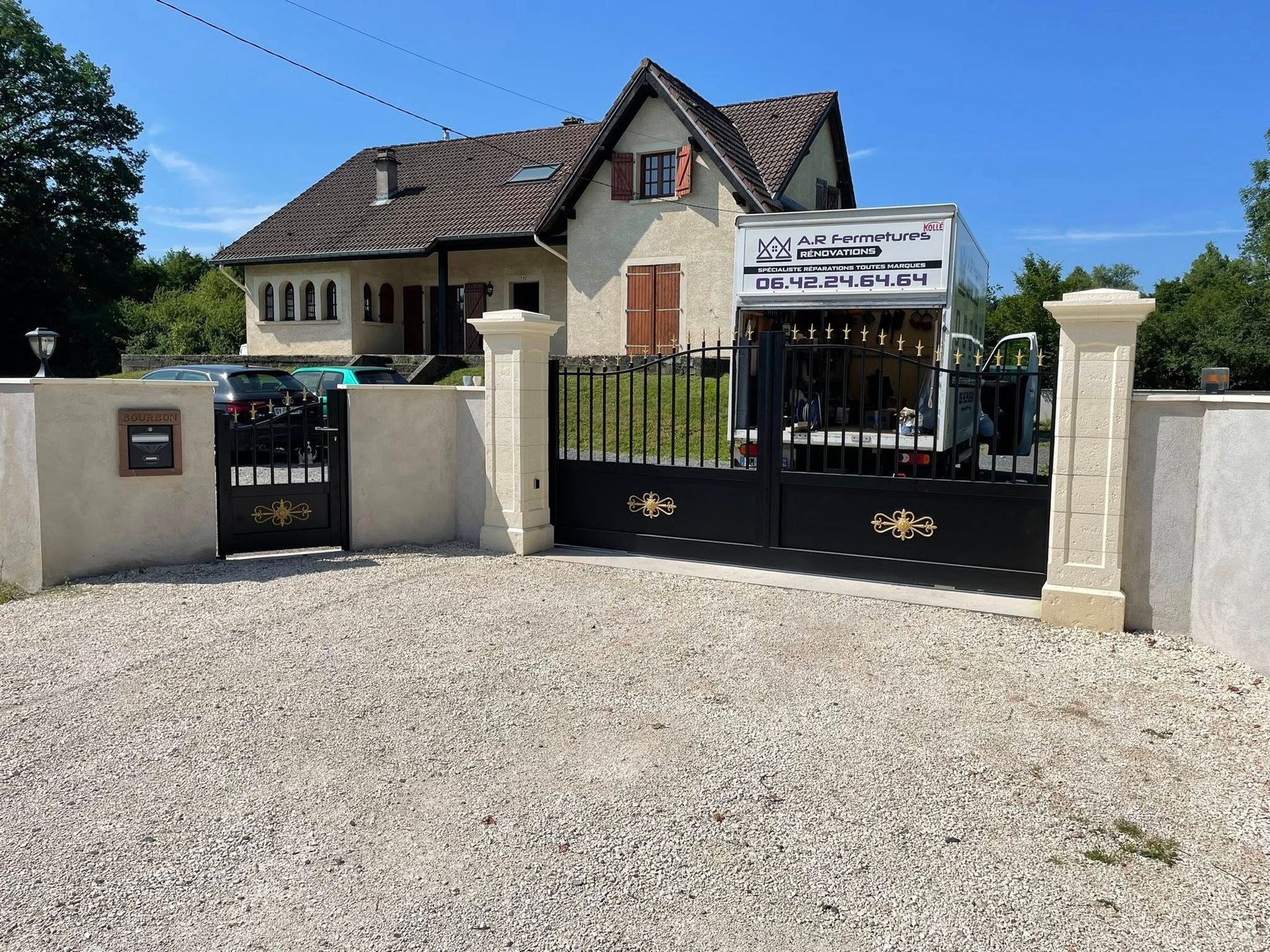 Maison beige avec portail noir, piliers et allée de gravier. Un camion blanc est garé derrière le portail.