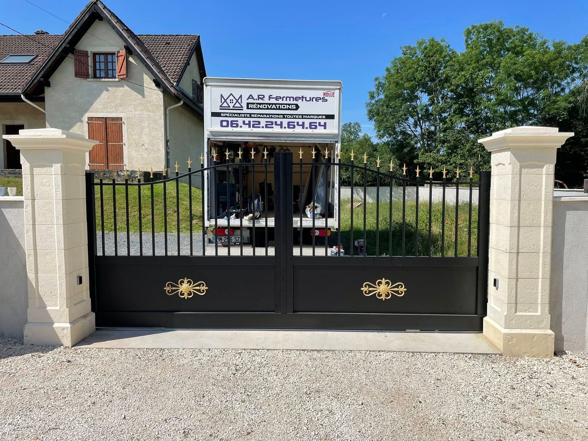 Portail en fer noir avec des accents dorés et des piliers en pierre beige, allée, camion de déménagement.
