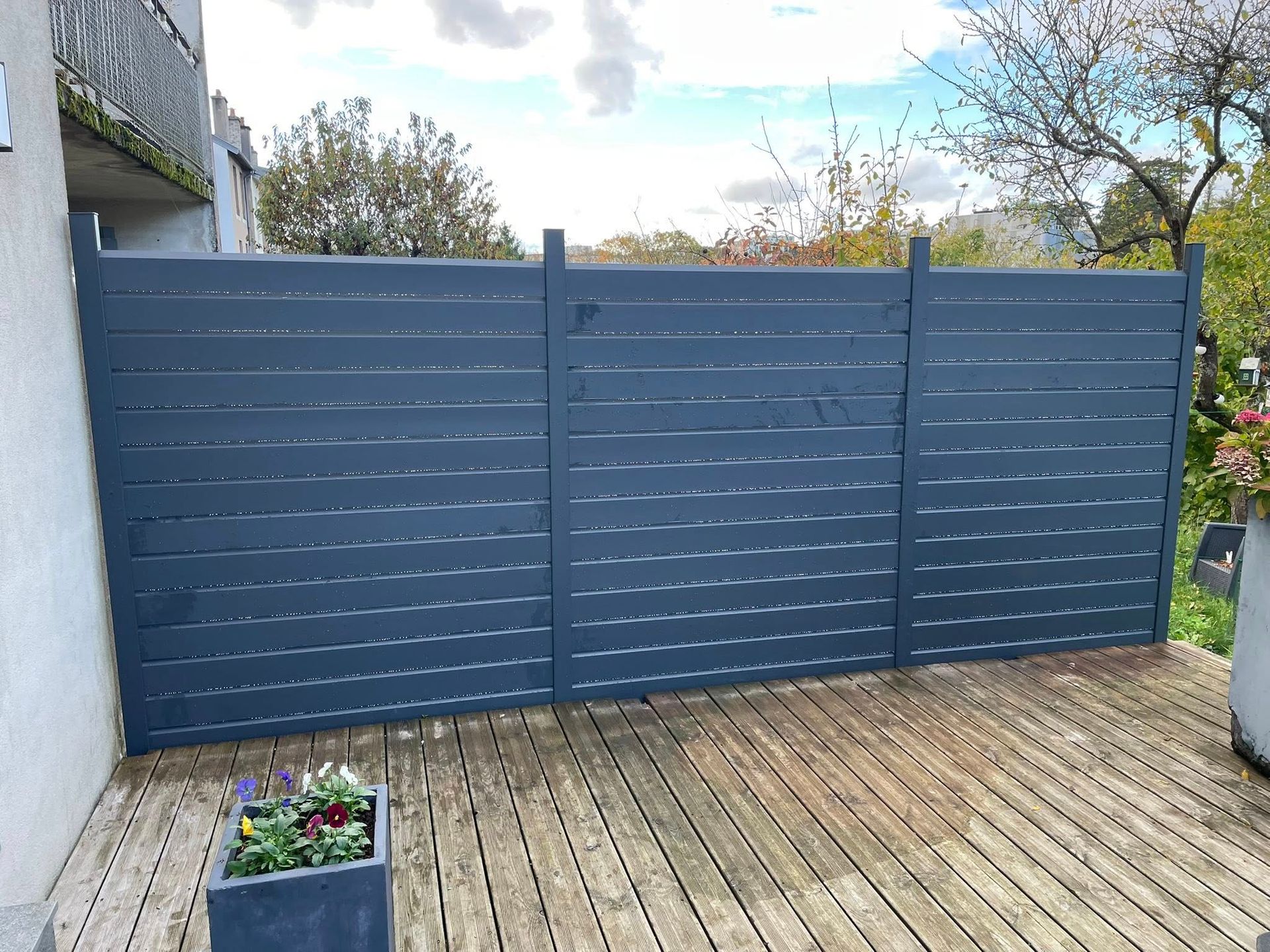 Une clôture en bois peinte en bleu préserve l'intimité d'une terrasse en bois. Une plante en pot est posée sur la terrasse, devant la clôture.