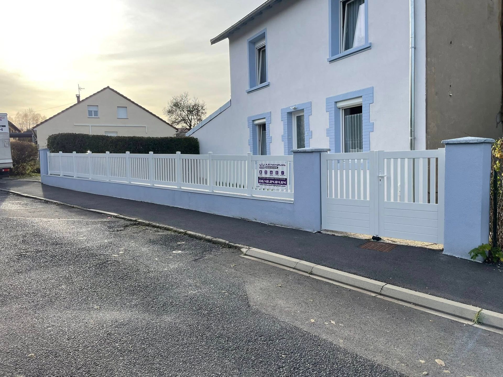 Clôture et portail blancs devant une maison bleu clair, au bord d'une rue.