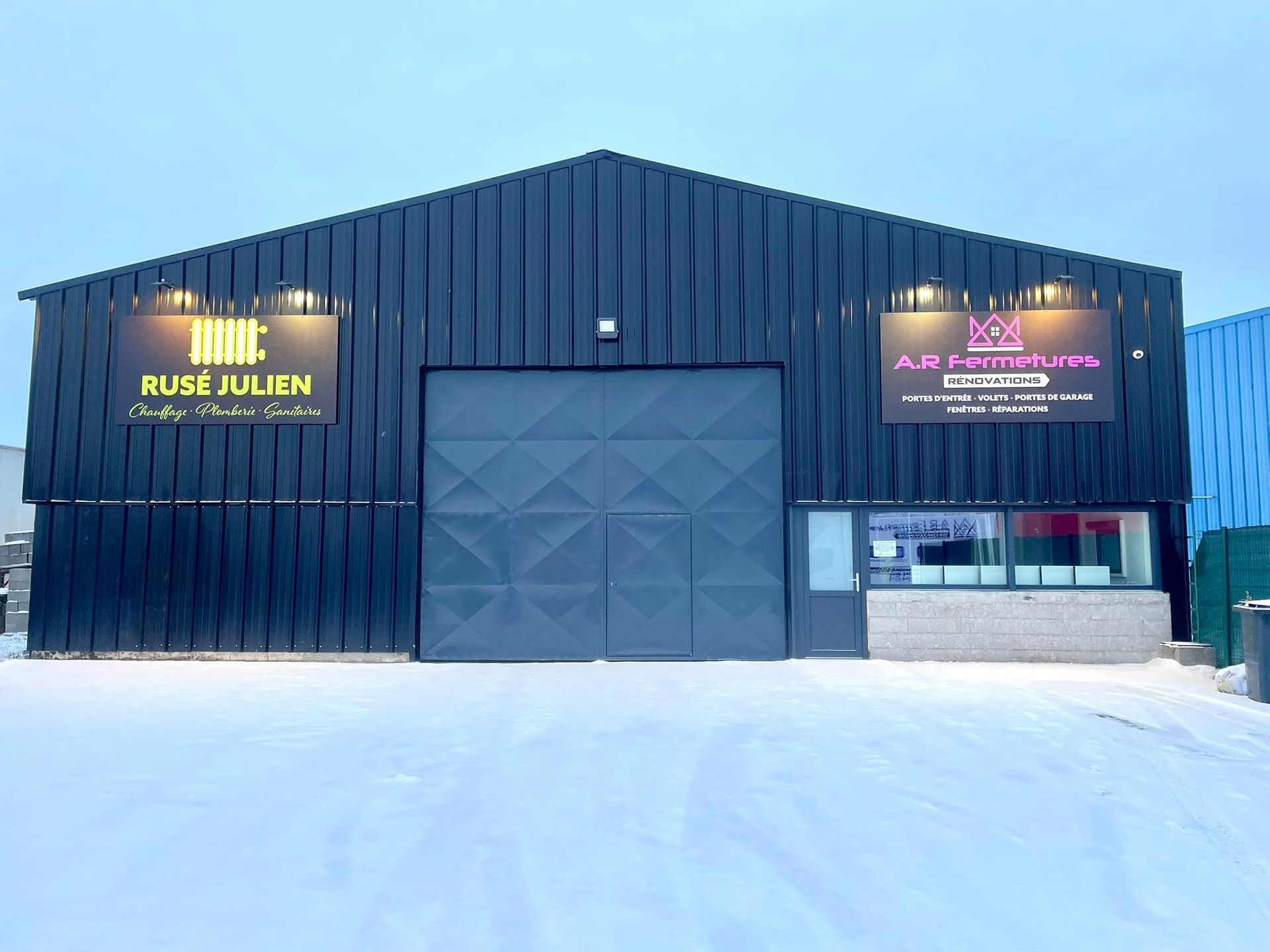 Bâtiment noir avec une grande porte, deux panneaux et une fenêtre. Sol enneigé devant.