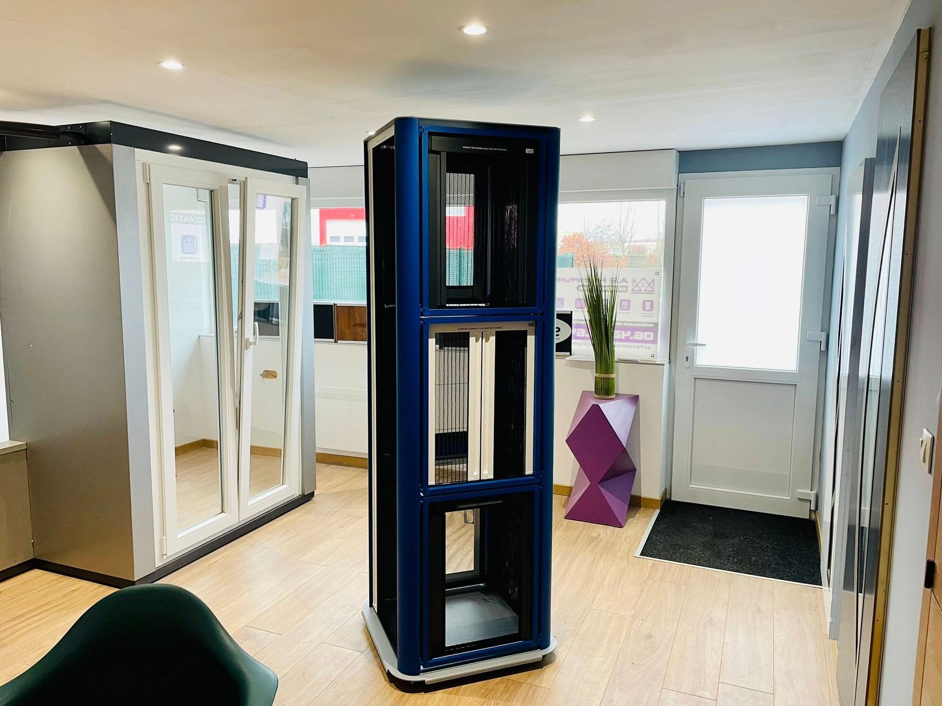 Vitrine bleue dans un showroom, présentant des exemples de portes et de fenêtres. Vue intérieure avec une porte, une plante et un parquet.