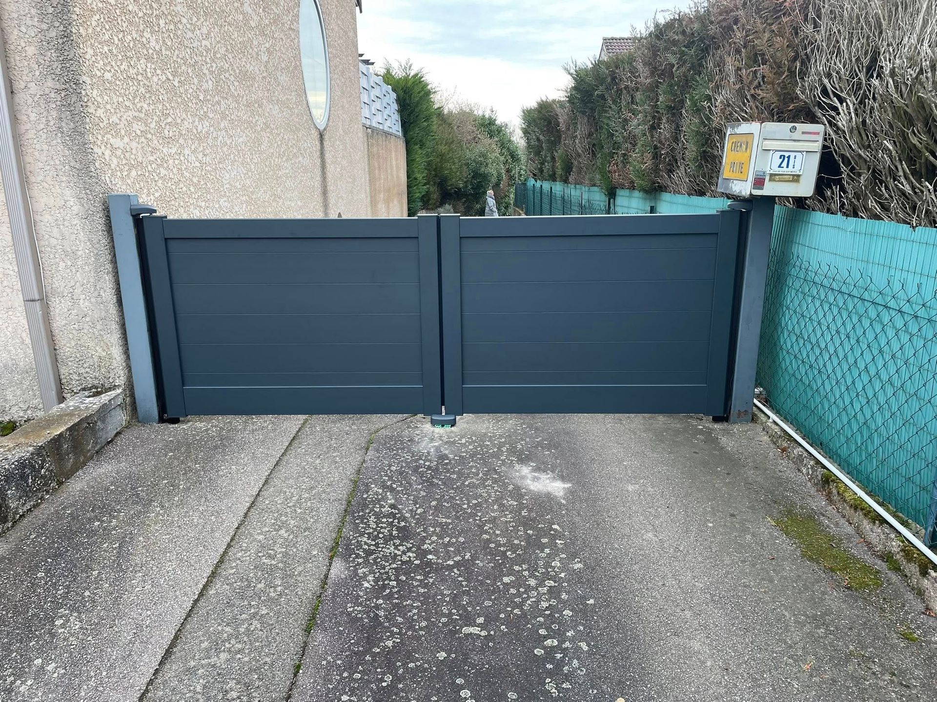 Portail gris à double battant barrant une allée. Lattes horizontales gris foncé, assise en béton et boîte aux lettres.