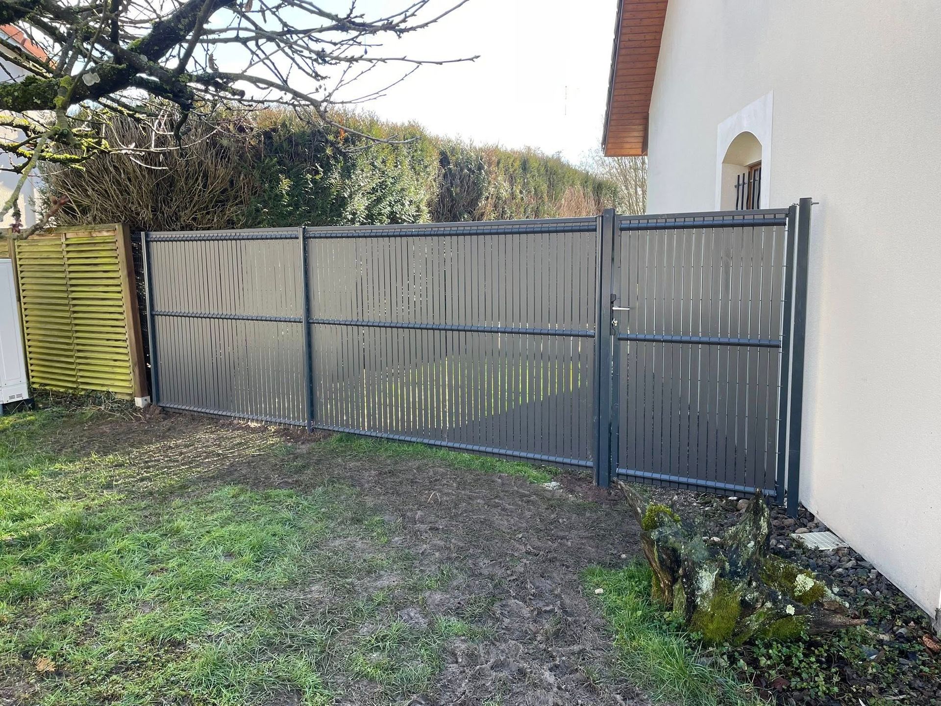 Clôture gris foncé avec portail à côté d'un bâtiment de couleur claire, pelouse verte et petits arbustes.