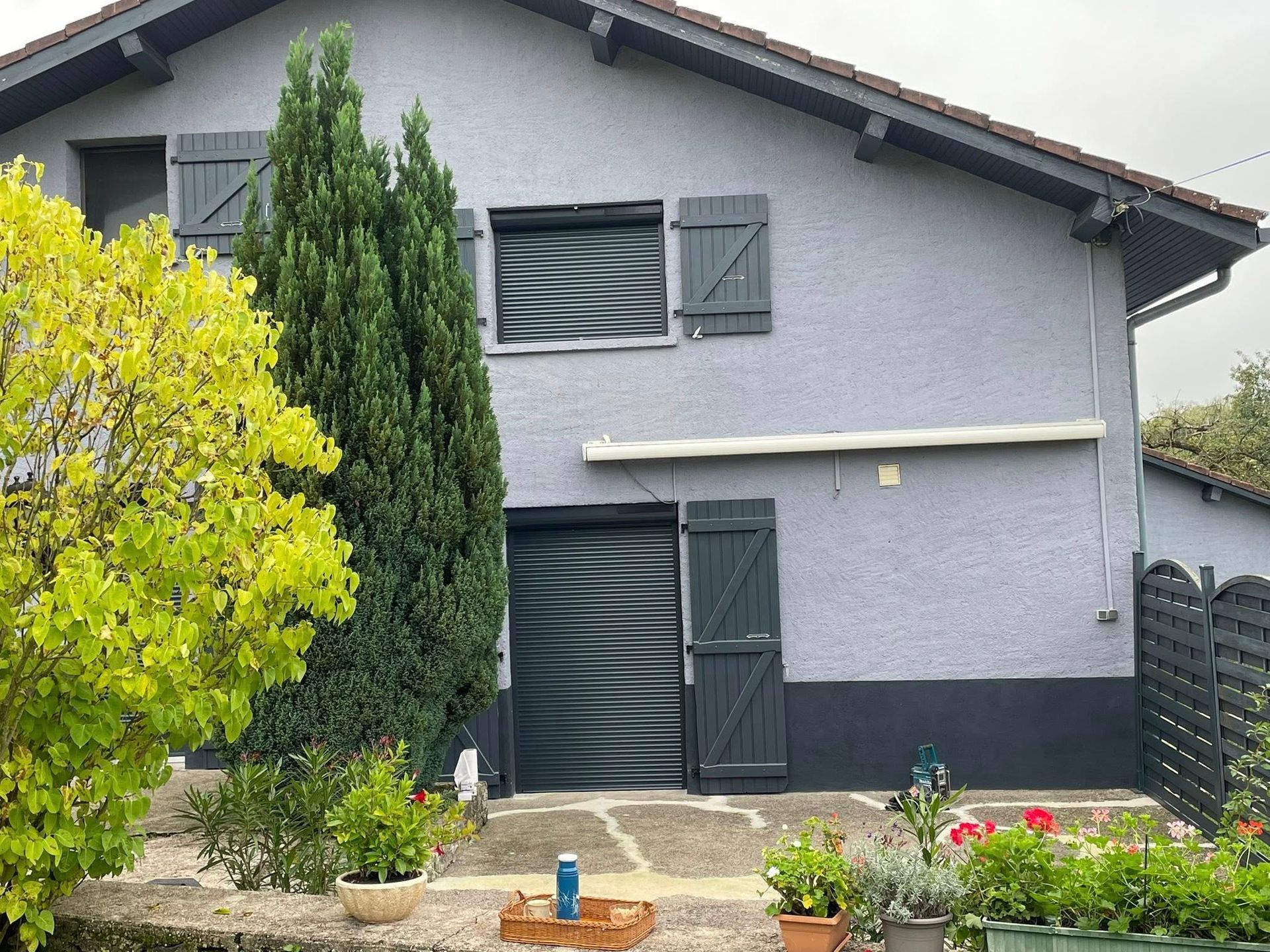 Maison grise aux volets sombres, grand arbre vert et jardin devant.