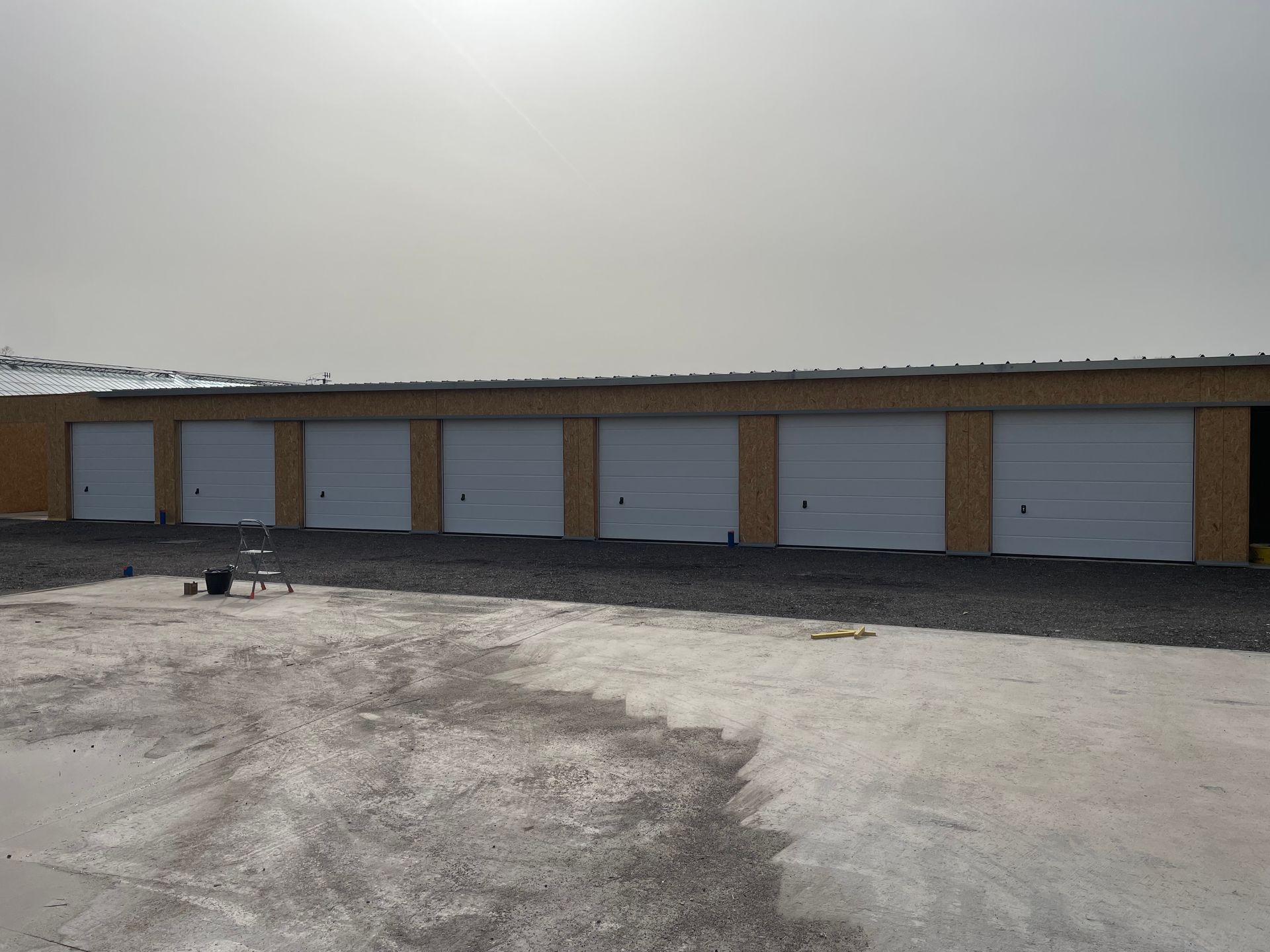 Des box de stockage aux portes blanches, aux murs beiges et un parking en gravier sous un ciel couvert.