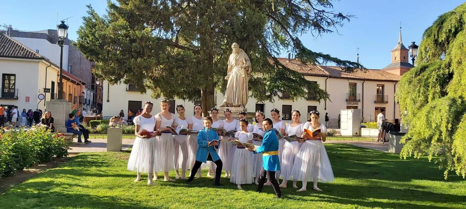 Un grupo de bailarines posa en un parque, con una estatua y edificios al fondo. Algunos llevan vestidos blancos, otros azules.