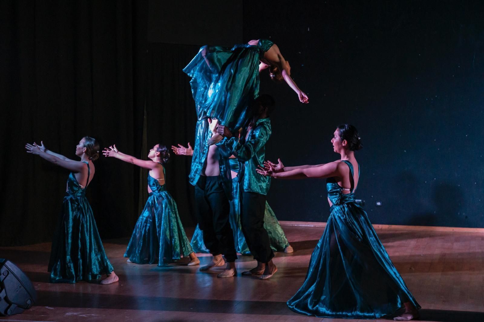 Bailarines con vestidos verde azulado levantan a una bailarina en el aire sobre un escenario. Escenario oscuro con iluminación teatral.