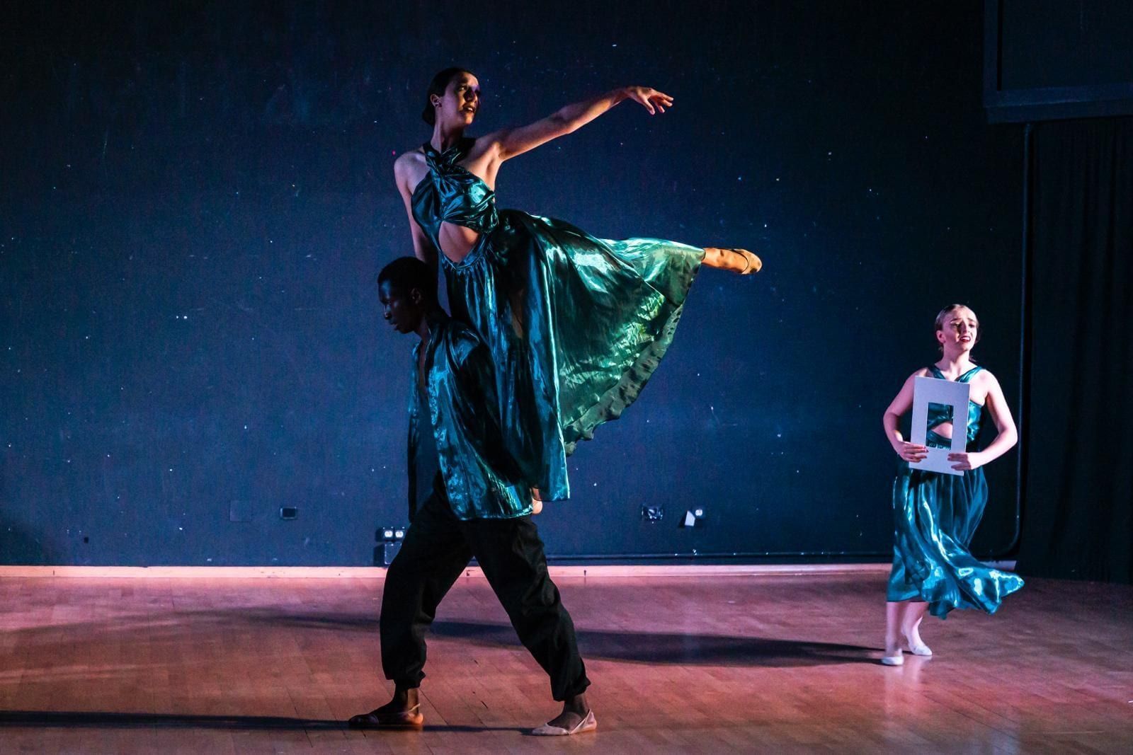 Bailarines con vestidos verde azulado en el escenario. Un hombre carga a una mujer. Un tercer bailarín sostiene una sábana.