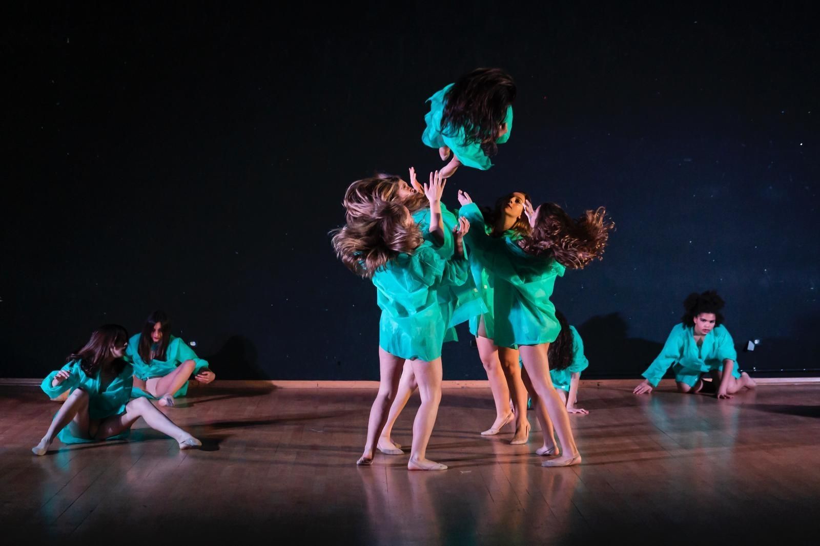 Bailarines con camisas verde azulado actúan en un escenario de madera con un fondo estrellado. Algunos estiran los brazos hacia arriba, otros hacia el suelo.