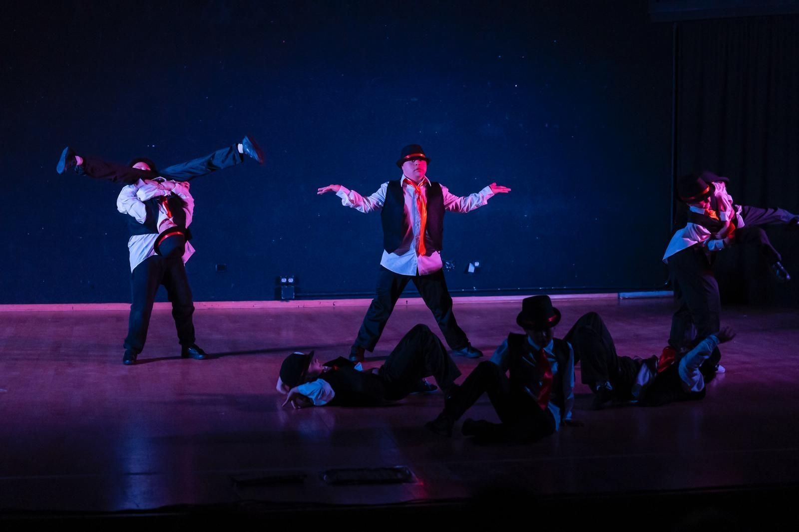 Un espectáculo de danza en el escenario. Cinco personas, una boca abajo, otras posando con los brazos en alto, de rodillas o de pie. Escenario oscuro.
