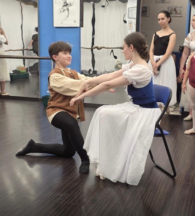 Niño arrodillado, con las manos extendidas hacia una mujer con falda blanca sentada en una silla, en un estudio de danza.