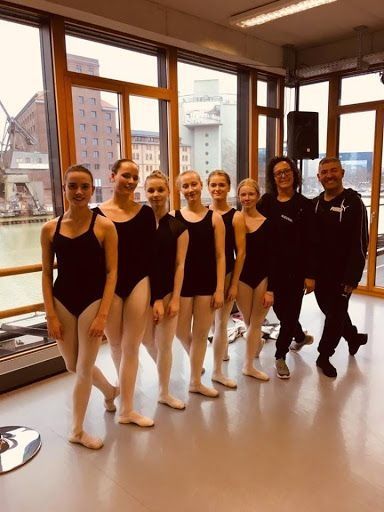 Bailarines de ballet y dos adultos posan en un estudio con grandes ventanales con vistas a un paisaje urbano.