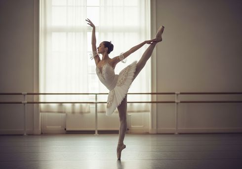 Bailarina con traje blanco, iluminada por un foco, en una pose dinámica, con los brazos extendidos, las rodillas dobladas y fondo negro.