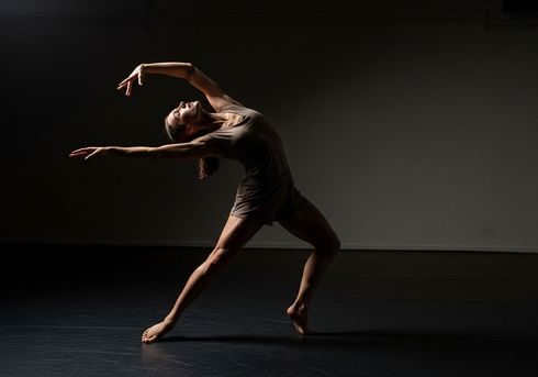 Bailarina con traje blanco, iluminada por un foco, en una pose dinámica, con los brazos extendidos, las rodillas dobladas y fondo negro.