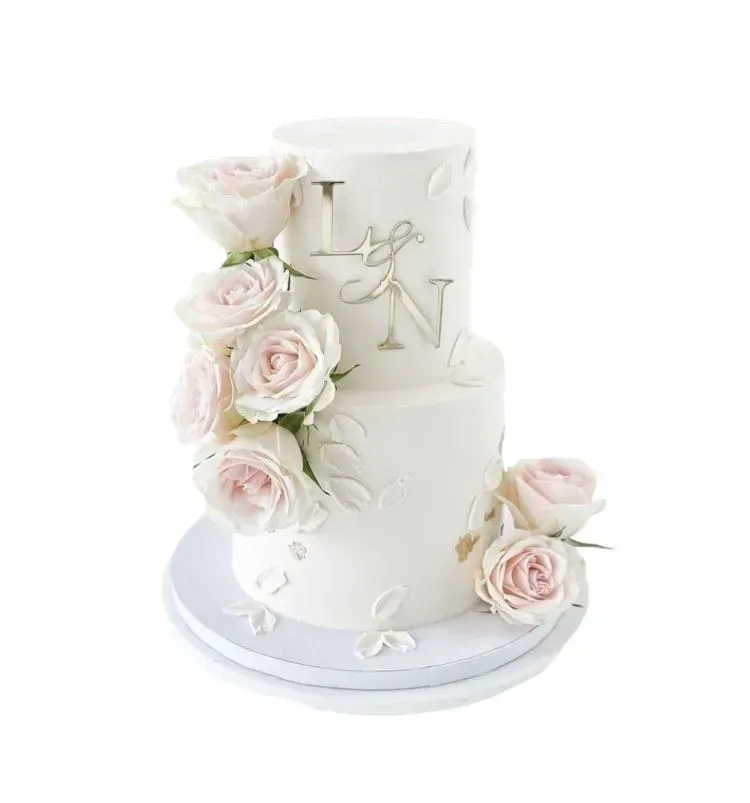 Tarta de boda blanca de dos pisos con rosas rosa pálido e iniciales doradas sobre un soporte blanco.