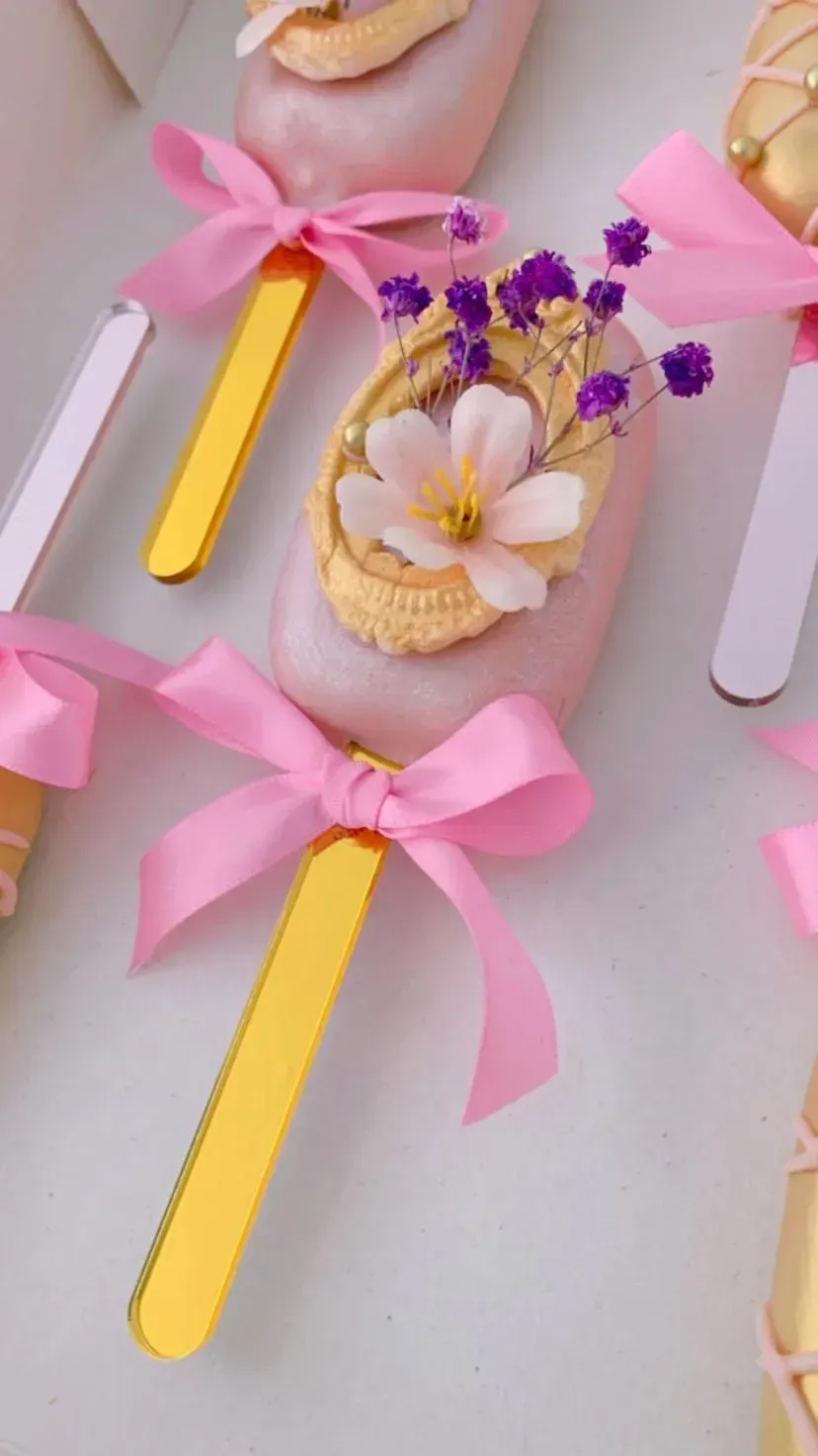 Palitos de manualidades amarillos con lazo rosa decorados con flores y una concha sobre un fondo pastel.
