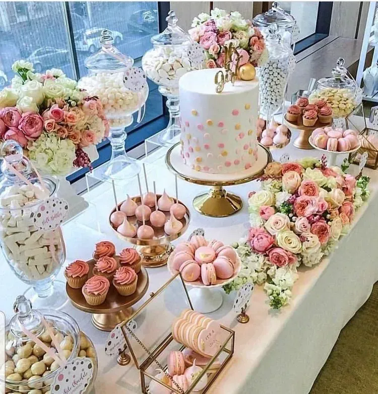Mesa de postres en tonos pastel con pastel, macarons y arreglos florales junto a una ventana.
