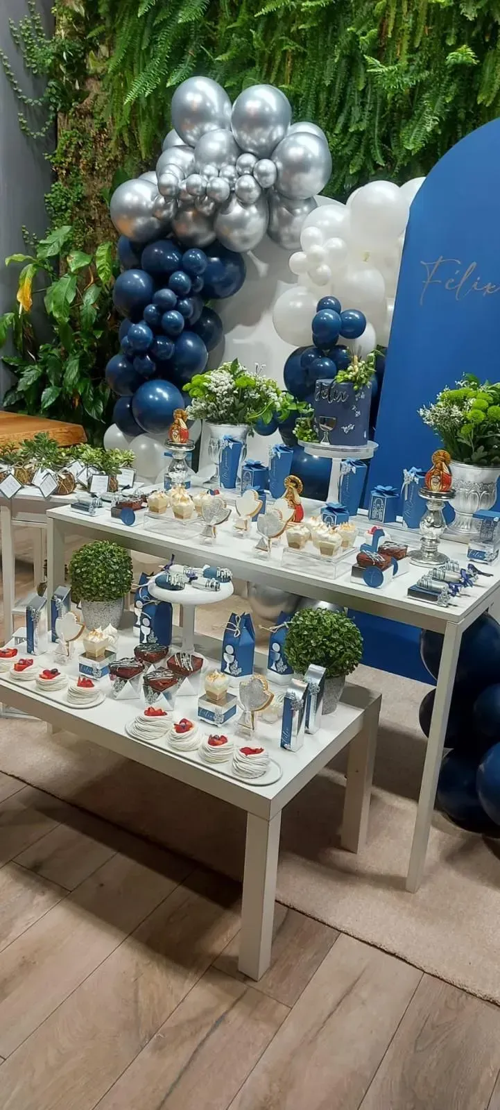 Decoración con globos azules y plateados detrás de una mesa de postres con pequeños pasteles y dulces.