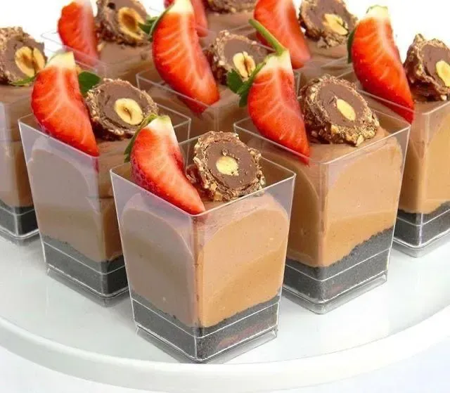 Vasitos de mousse de chocolate cubiertos con rodajas de fresa, avellanas y galletas de chocolate sobre un plato blanco.