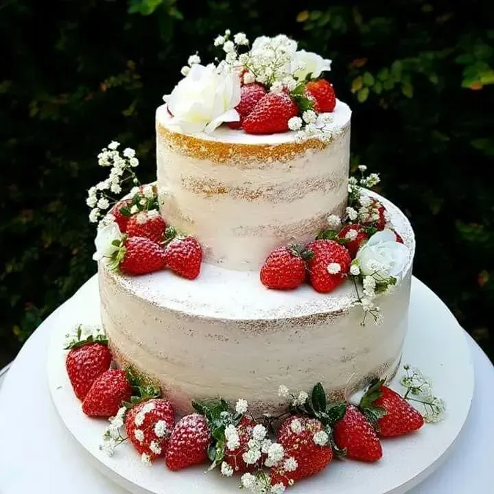 Tarta desnuda de dos pisos con fresas, nata montada y paniculata sobre un soporte blanco.