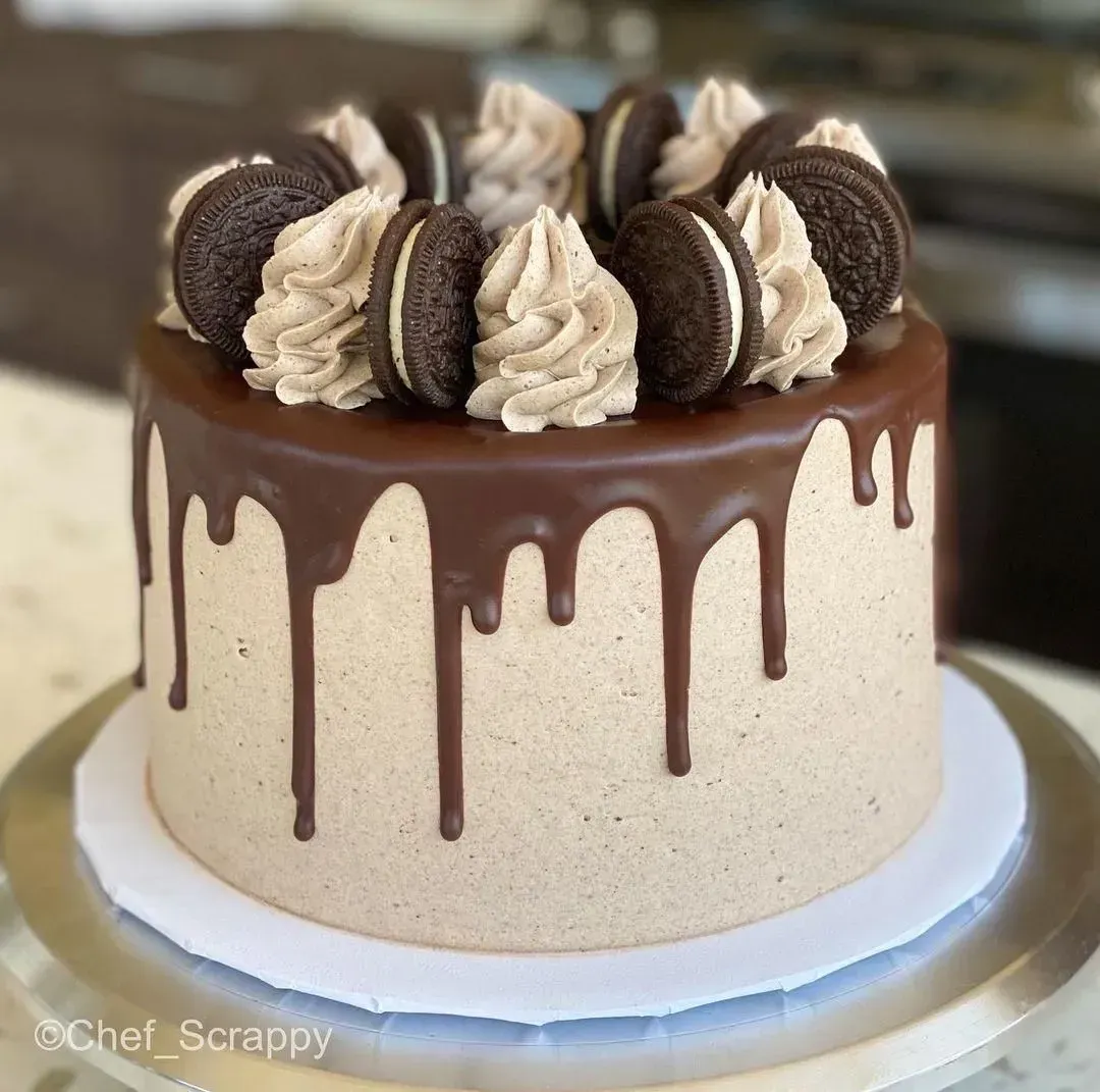 Pastel de chocolate con glaseado chorreante, decorado con espirales de crema y galletas Oreo sobre un soporte blanco.
