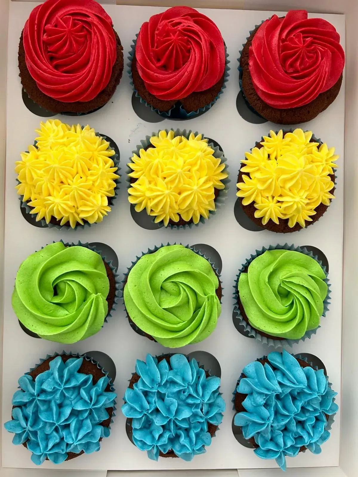 Filas de cupcakes rojos, amarillos, verdes y azules en una cuadrícula de 4x3 sobre una bandeja.