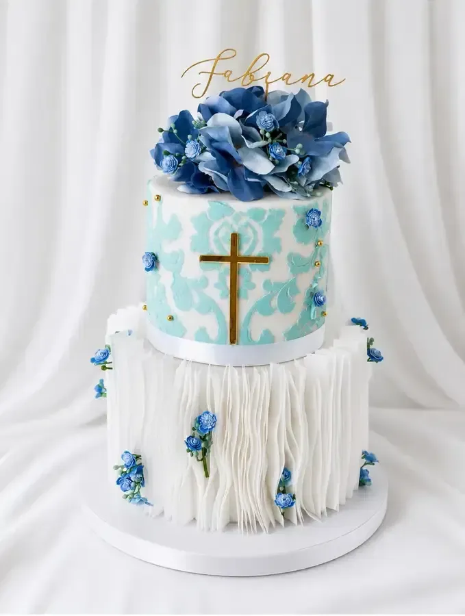 Tarta blanca de dos pisos con flores azules, cruz dorada y adorno con letras doradas sobre una base blanca.