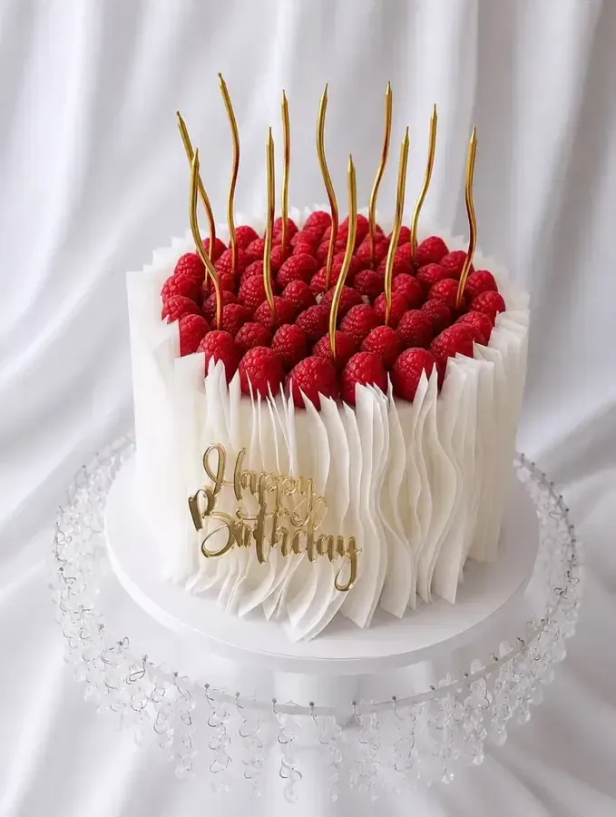 Pastel blanco con frutos rojos y velas doradas, decorado con glaseado blanco sobre un soporte de encaje.
