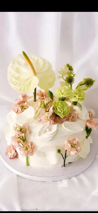 Tarta blanca con flores rosas, detalles verdes y un adorno de anturio color crema sobre una base blanca.