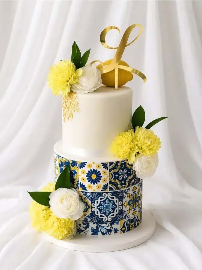 Tarta floral de dos pisos en blanco y azul con flores amarillas y adorno dorado sobre un fondo blanco drapeado.