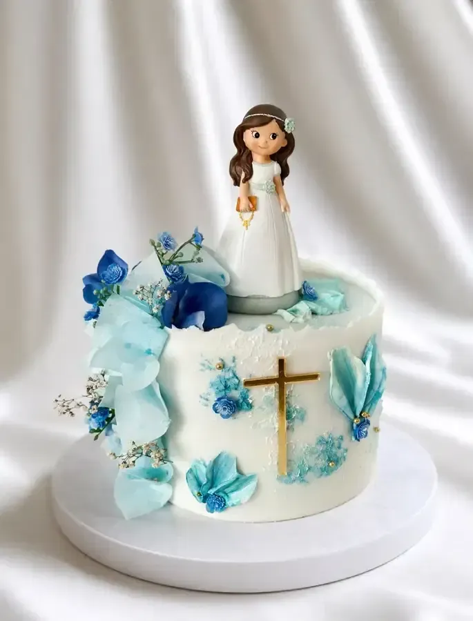 Pastel blanco con flores azules, una cruz dorada y una figurita de una niña pequeña encima, sobre un mantel blanco.