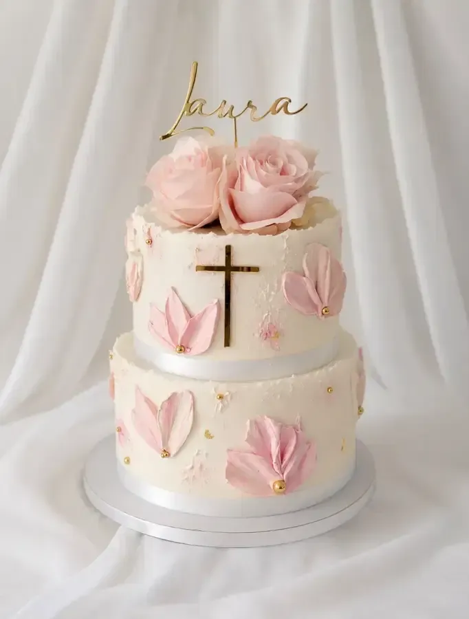 Tarta blanca de dos pisos con rosas rosadas, cruz dorada y adorno dorado con el nombre sobre una base blanca.