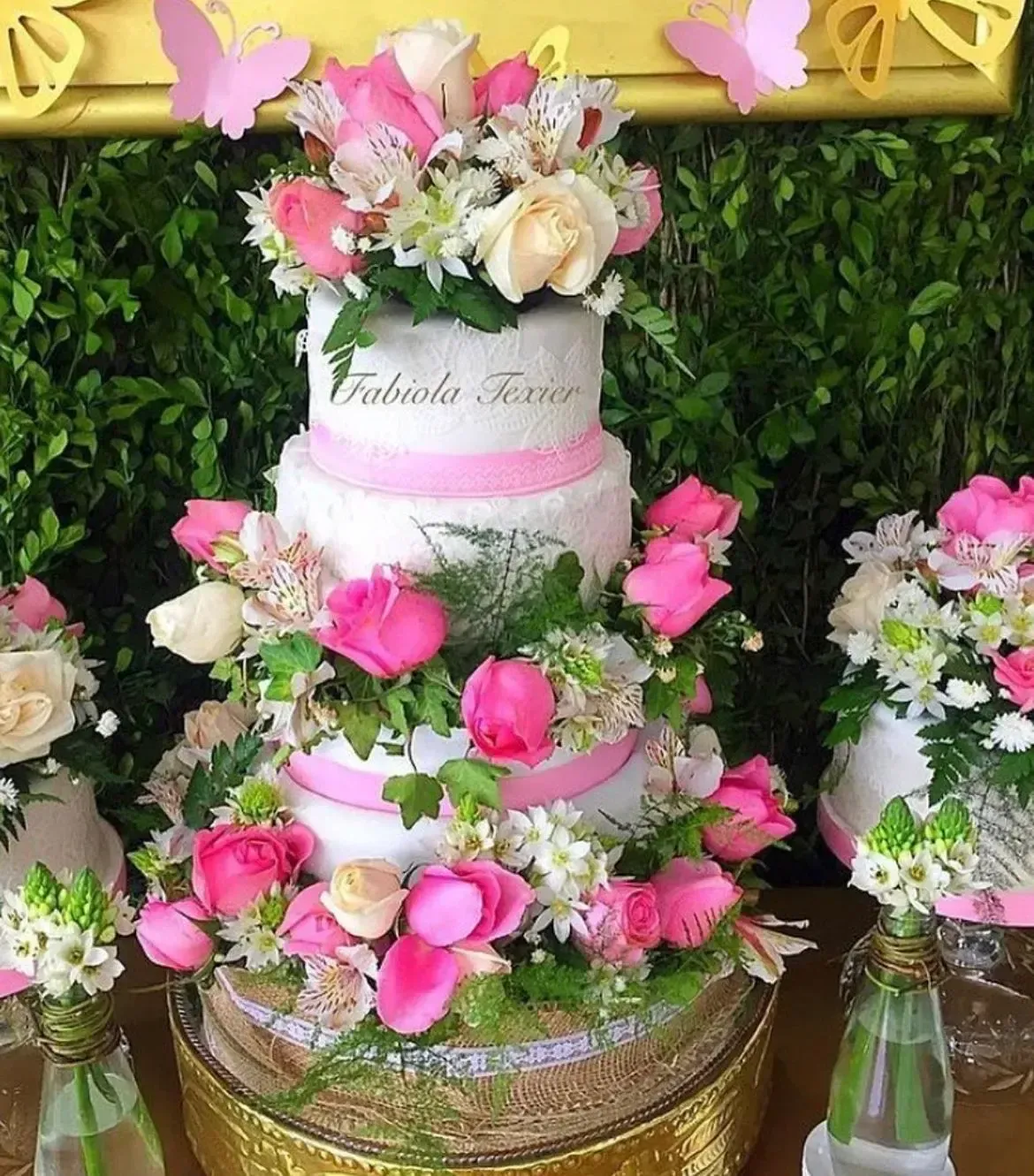 Tarta blanca de tres pisos con flores rosas sobre una base dorada, expuesta sobre un fondo verde.